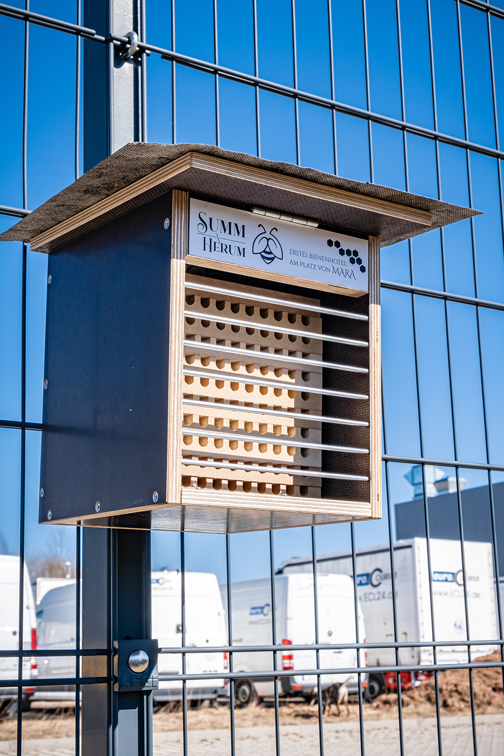 Bienenhotel am Zaun, im Hintergrund Kurierfahrzeuge, Kleintransporter und LKW.